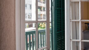 Une vue à travers une fenêtre ornée d’un volet vert, avec une porte blanche et un balcon doté d’une balustrade blanche.