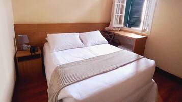 Un lit bien fait, avec du linge de lit blanc, une tête de lit en bois, une table de chevet avec une lampe et une fenêtre avec des volets.