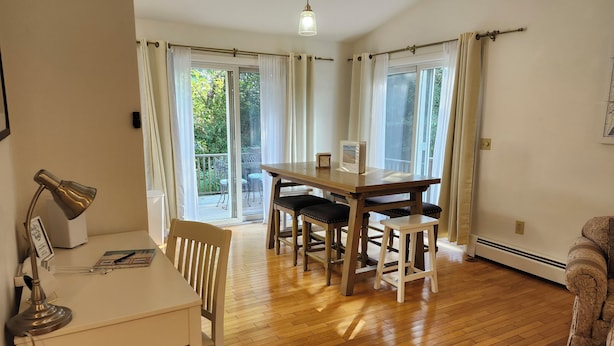 Un coin repas avec une table et des chaises en bois, un bureau blanc avec une lampe, et une vue sur l’extérieur grâce à de grandes fenêtres.