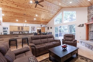 A spacious living area with a wooden ceiling, a large sofa, a coffee table, and a fireplace. - Single Level, Firepit + Large Deck, Game Room (Lake Arrowhead)