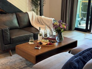 A modern living room with a leather sofa, a wooden coffee table with snacks and drinks, a vase with flowers, and a view through a glass door. - Summer Adventure Awaits | Modern Cottage - 2 (Sevierville)