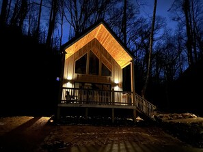 A wooden cabin with a porch and a large window, surrounded by trees.