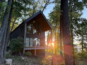 Une cabane dotée d’un balcon, entourée d’arbres, avec un coucher de soleil en arrière-plan.