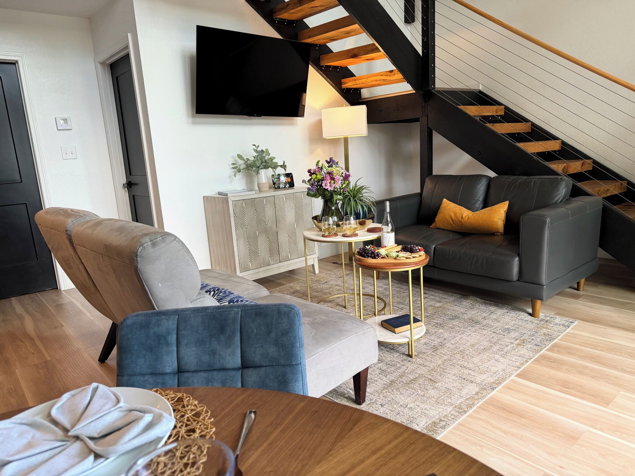 A modern living room with a sofa, a dining table, a TV mounted on the wall, and a staircase.