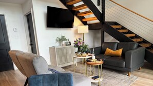A modern living room with a sofa, a dining table, a TV mounted on the wall, and a staircase. - One Last Summer Getaway -1 (Sevierville)