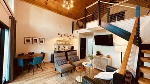 A modern loft-style living space with a wooden ceiling, a dining area with a round table and blue chairs, a seating area with a sofa and armchairs, a kitchen with a built-in island, and a staircase leading to an upper level. - One Last Summer Getaway -1 (Sevierville)