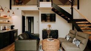 Un salon moderne avec un canapé, un fauteuil et une table basse ronde. On y trouve un escalier avec une rampe en bois et une balustrade d’escalier en métal noir.