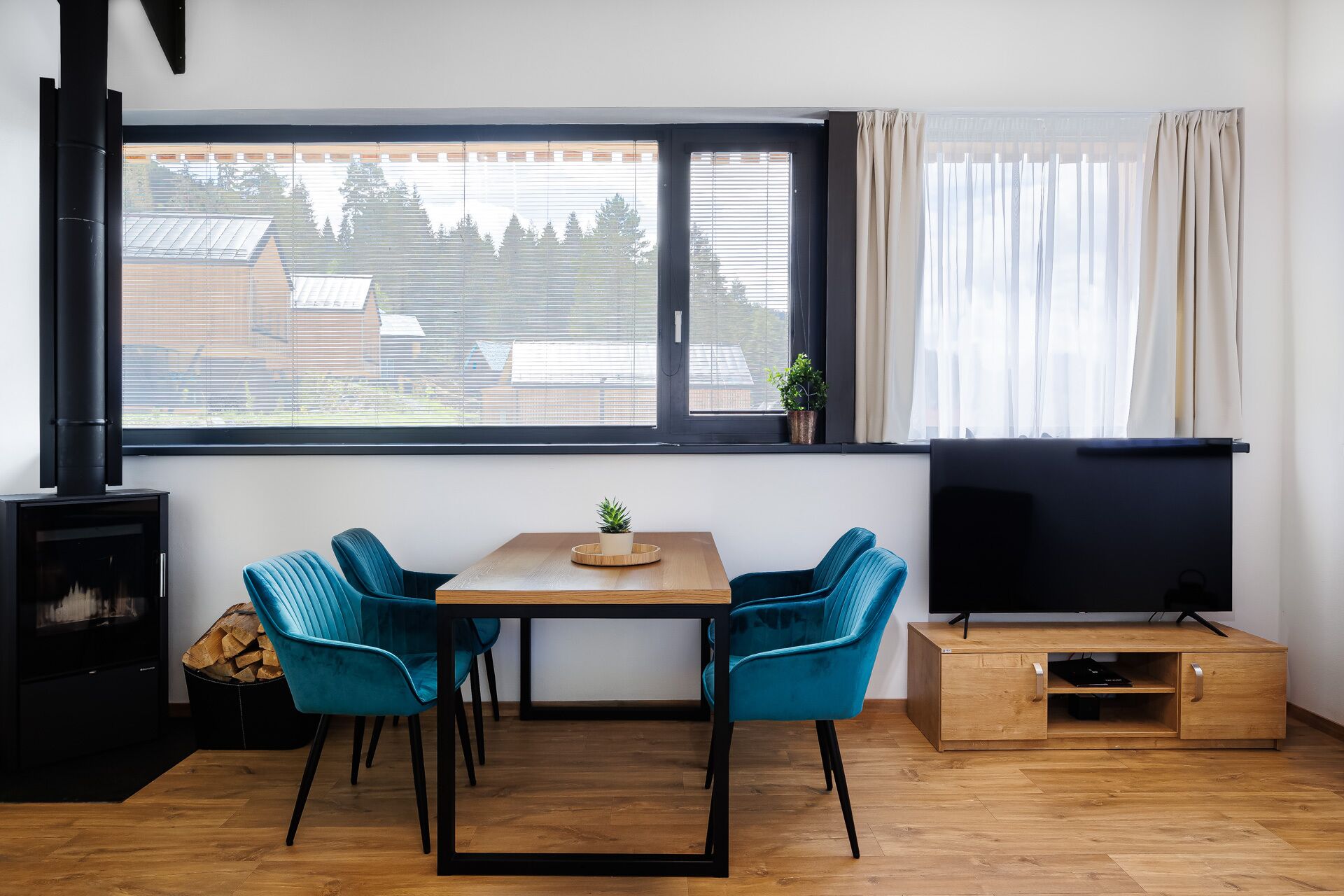 A modern living room with a wooden table, two blue chairs, a TV on a wooden stand, a fireplace, and a large window with blinds.