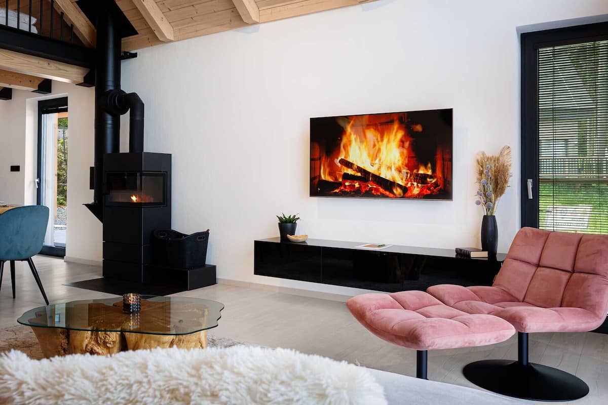 A modern living room with a black wood-burning stove, a glass coffee table, a pink upholstered chair, and a flat-screen TV displaying a fire.