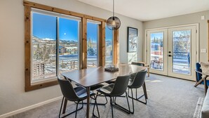 Un comedor con una mesa grande y cuatro sillas, ventanas amplias con vistas a un paisaje nevado y una lámpara colgante.