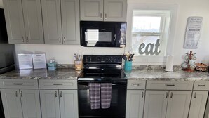 A kitchen with grey cabinets, a black stove, a microwave, and a window with the word "eat" on it. There are signs on the counter and a dish rack with cups. - 5-Mins to Cedar Point, 4-bedroom house. (Sandusky)