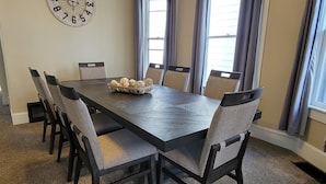 A dining room with a long table, six chairs, a clock on the wall, and a window with curtains. - 5-Mins to Cedar Point, 4-bedroom house. (Sandusky)