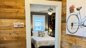 A bedroom with a wooden floor, a bed with white bedding, a nightstand with a lamp, a window with blinds, and a painting of a bicycle on the wall. - Charming Bungalow On Main St & the Swamp Rabbit Trail with Private Pool (Travelers Rest)