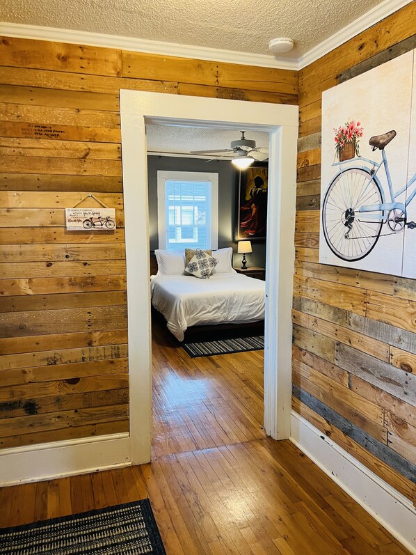 A bedroom with a wooden floor, a bed with white bedding, a nightstand with a lamp, a window with blinds, and a painting of a bicycle on the wall. - Charming Bungalow On Main St & the Swamp Rabbit Trail with Private Pool (Travelers Rest)