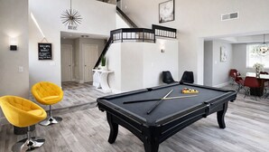 A modern living room with a pool table, two yellow chairs, and a staircase leading to an upper level.