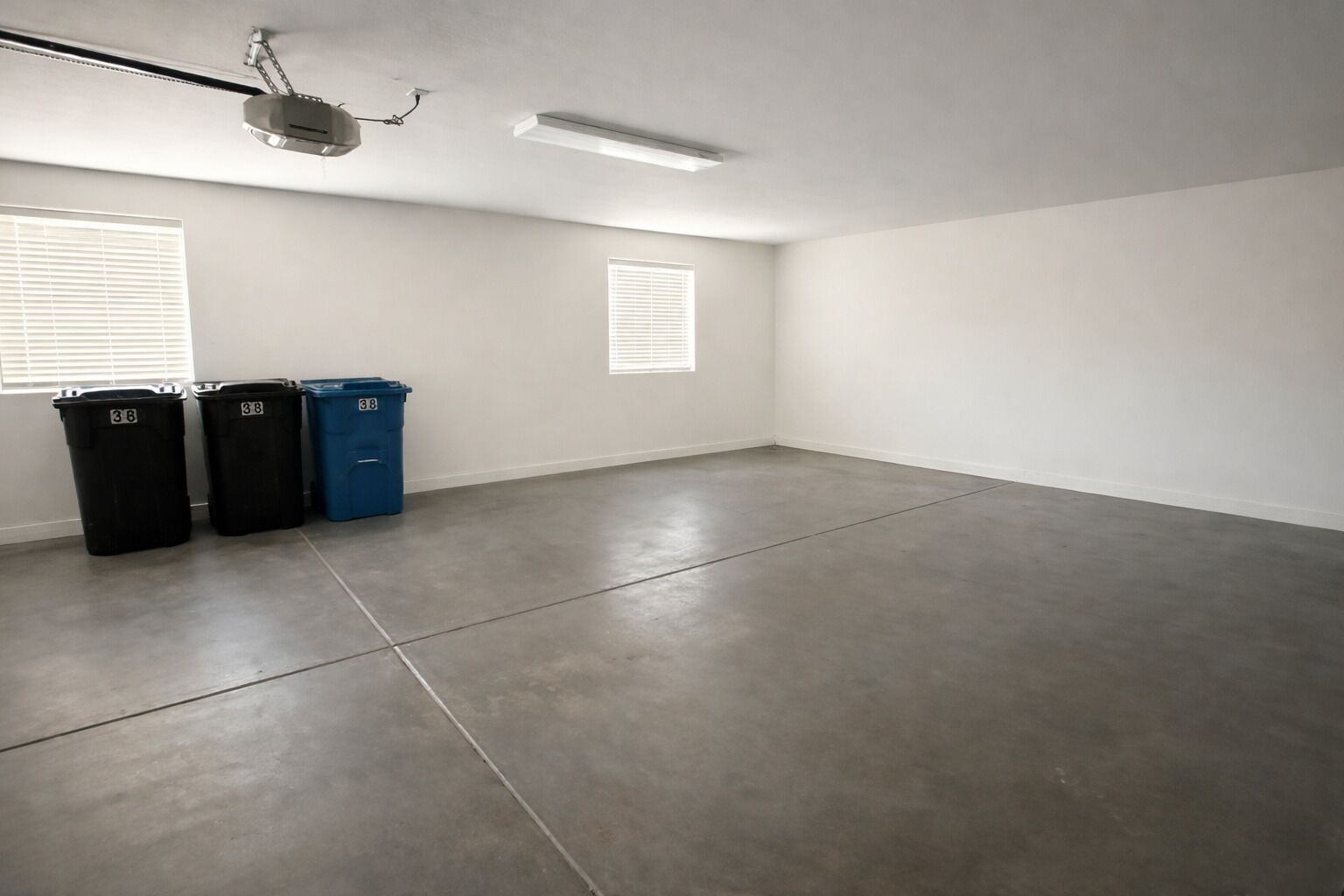 A spacious, empty room with a concrete floor, two windows with blinds, and three trash bins labeled '38'.