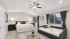 A bedroom with a bed, a sofa, a round table, a ceiling fan, a clock, and a window with curtains.