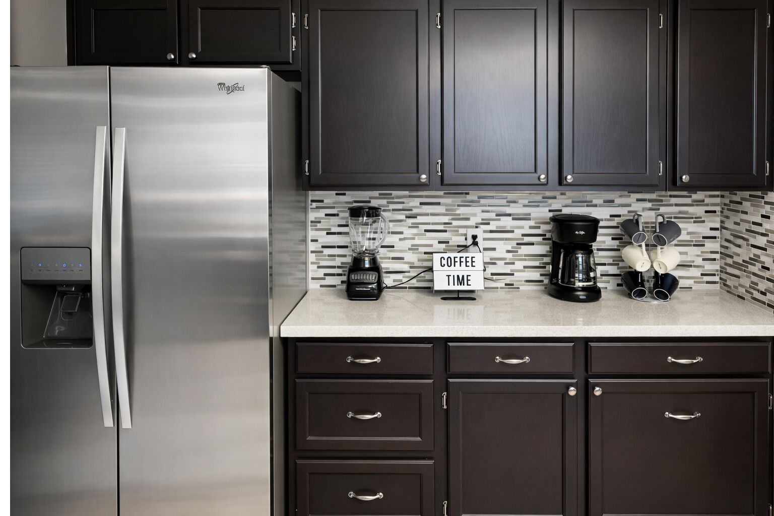 A modern kitchen with dark cabinets, a stainless steel refrigerator, a coffee maker, and a blender.