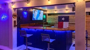 A bar area with a wooden counter, two bar stools, a television screen, and a Texas flag.