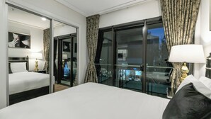 Hotellihuone, jossa on sänky, yöpöytä, lamppu, peili ja näkymä lasiovesta.