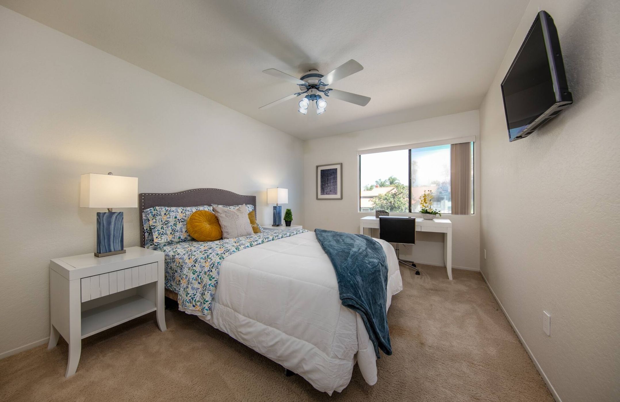 A bedroom with a bed, bedside tables, a desk, a ceiling fan, a TV, and a window.