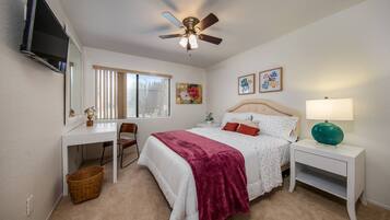 A bedroom with a bed, a desk, a chair, a ceiling fan, a TV, and artwork on the wall.