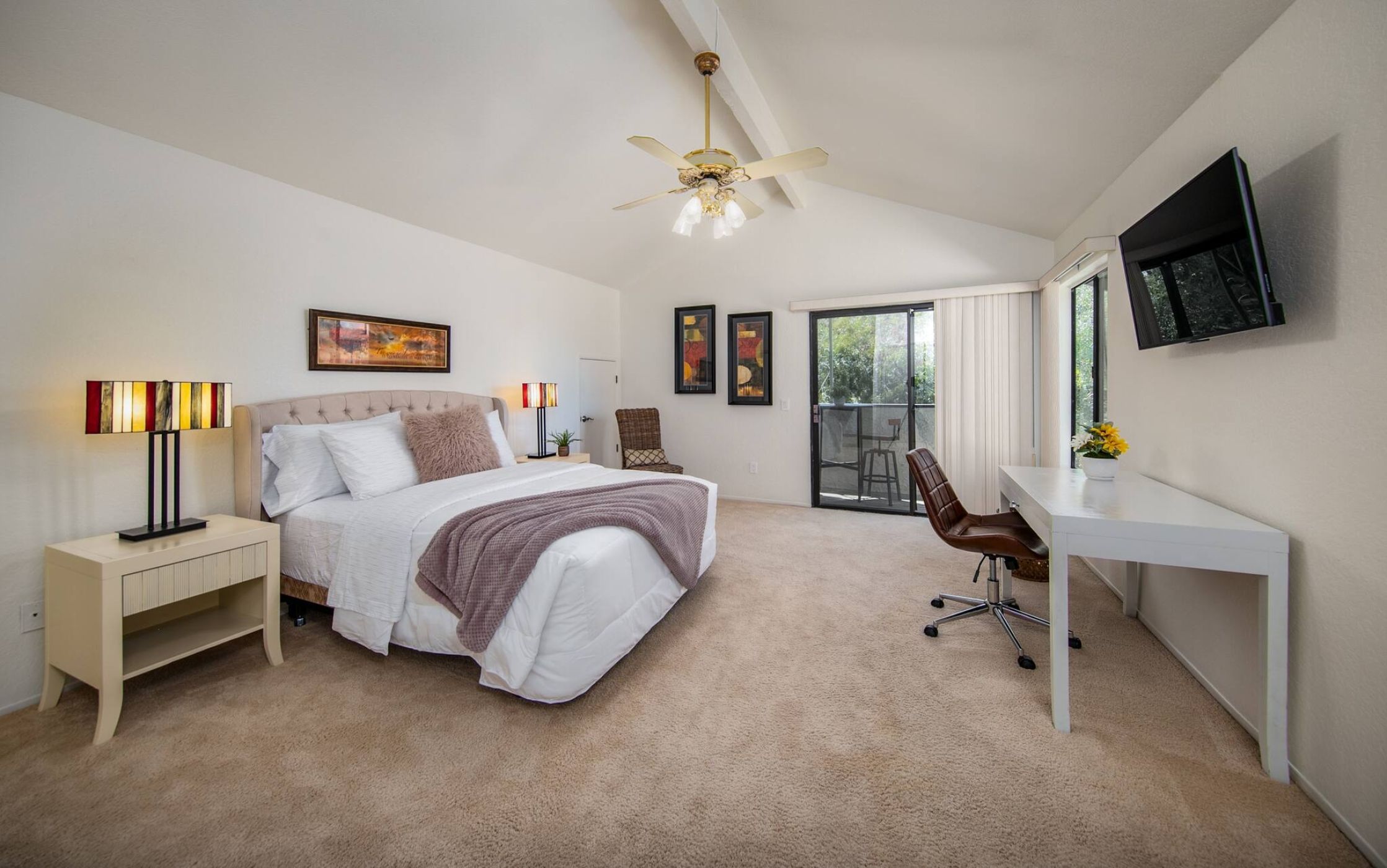 A bedroom with a bed, bedside tables, a desk, a chair, a ceiling fan, and a TV.