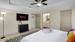 Ein Schlafzimmer mit einem Bett, einem Deckenventilator, einem an der Wand montierten Fernseher, einer Kommode und einer Tür zu einem Badezimmer.
