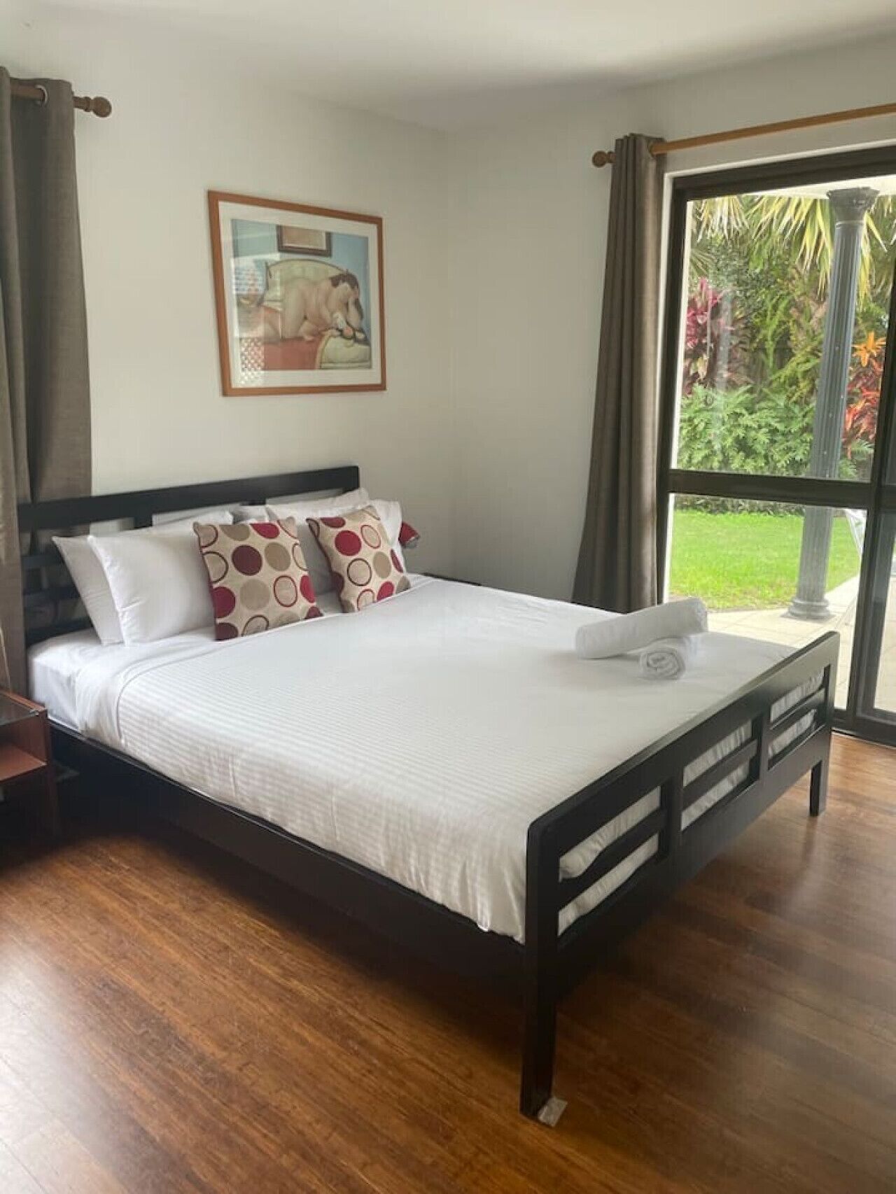 Una cama bien hecha con ropa blanca y almohadas estampadas, un cabecero de madera y un cuadro en la pared.