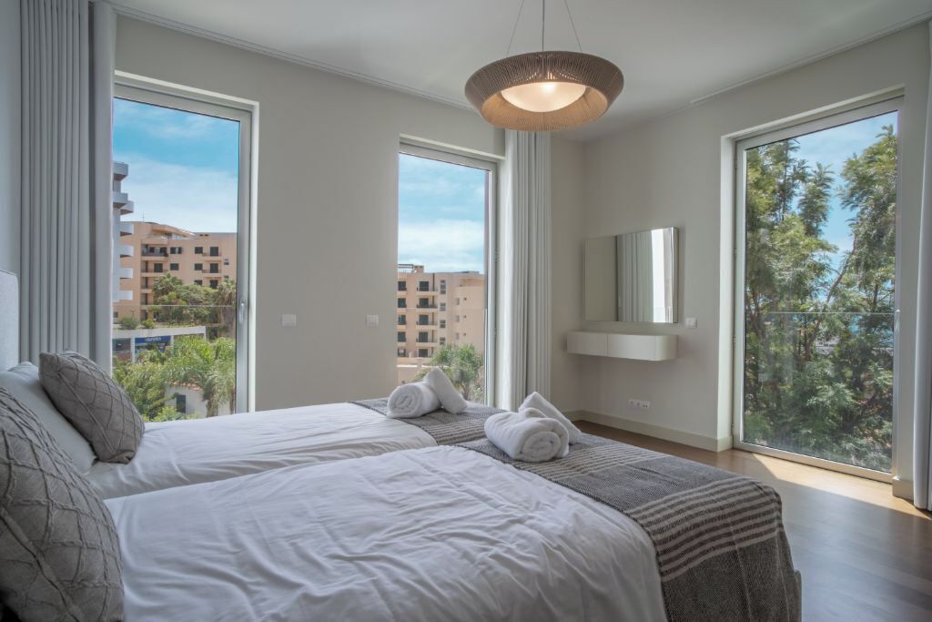 A modern hotel room with two beds, large windows, and a view of buildings and trees.