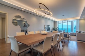 Un comedor moderno con una mesa de madera y sillas blancas, un espejo grande y vista al mar a través de amplias ventanas.