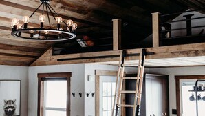 A loft-style room with exposed wooden beams, a chandelier with multiple bulbs, a ladder, and a rustic aesthetic.