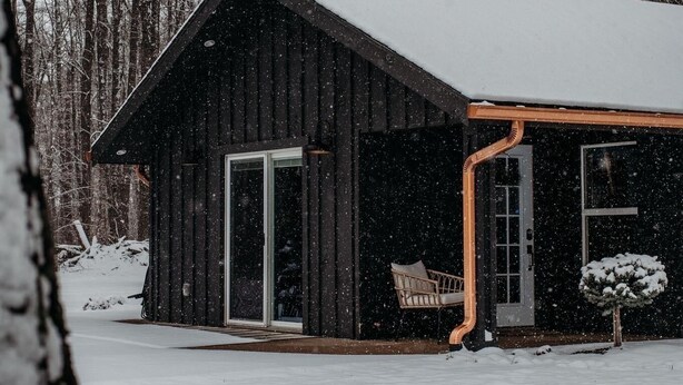 A wooden cabin with a snow-covered roof, large sliding glass doors, and a porch with a chair.
