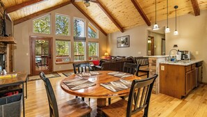 Un vaste espace de salle à manger avec une table et des chaises en bois, une cuisine dotée d’un îlot central en pierre et de grandes fenêtres offrant une vue sur l’extérieur.