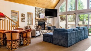 A spacious living room with a stone fireplace, a large blue sofa, and a wooden staircase. - Dog Friendly w/Guesthouse, Deck w/Grill, Foosball, Trail Access, Kid's Playhouse (Angel Fire)