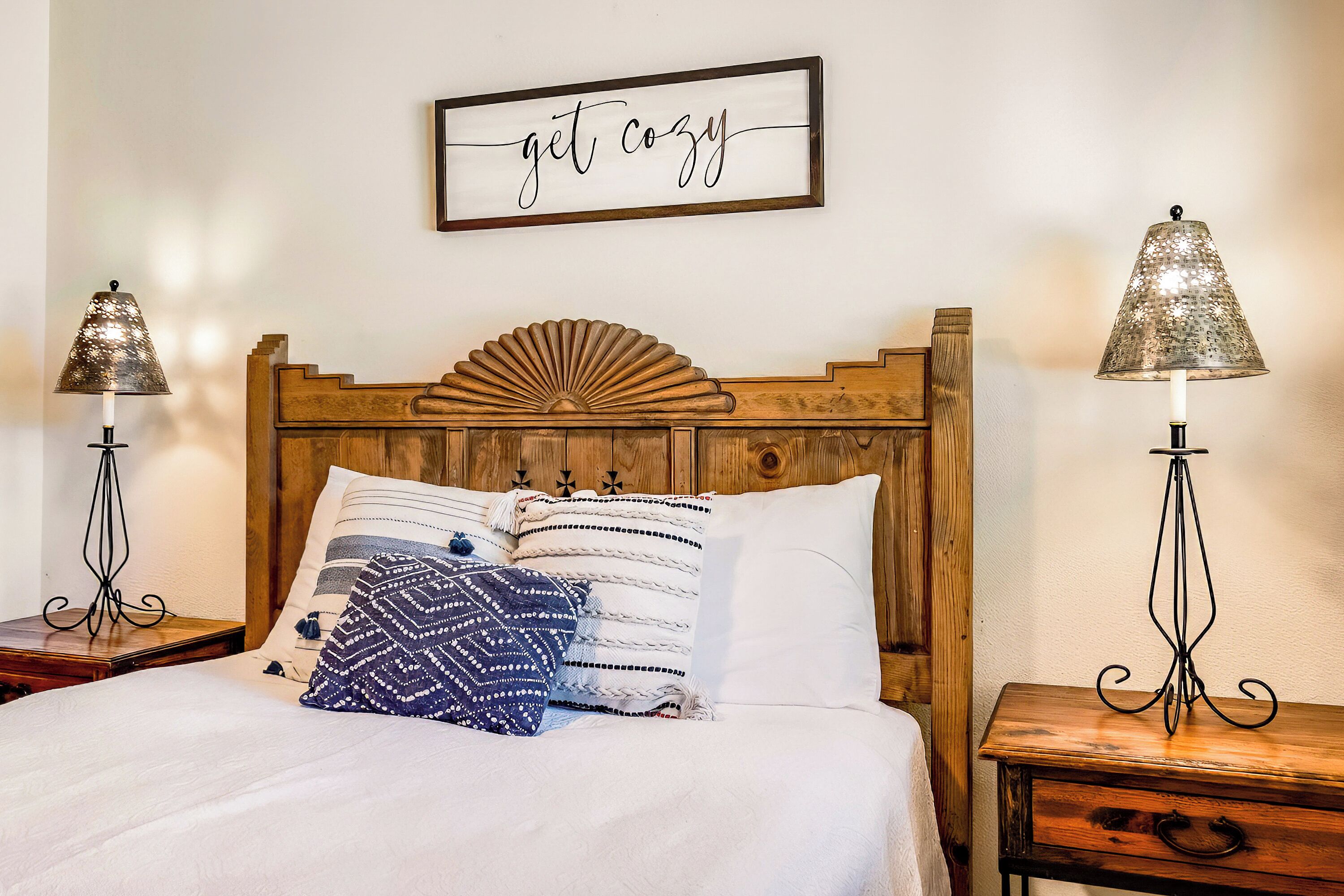 Ein ordentlich bezogenes Bett mit einem Holz-Kopfteil, zwei Nachttische mit Lampen und einem Wandschild mit der Aufschrift "get cozy".