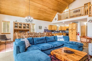 A spacious living room with a blue sectional sofa, wooden dining table, and a kitchen area with wooden cabinets.