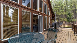 A wooden deck with a round metal table and chairs, large windows with blinds, and a view of a forest. - Dog Friendly w/Guesthouse, Deck w/Grill, Foosball, Trail Access, Kid's Playhouse (Angel Fire)