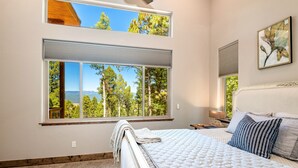 Un dormitorio con un ventanal grande, una cama, una mesita de noche y un cuadro en la pared.