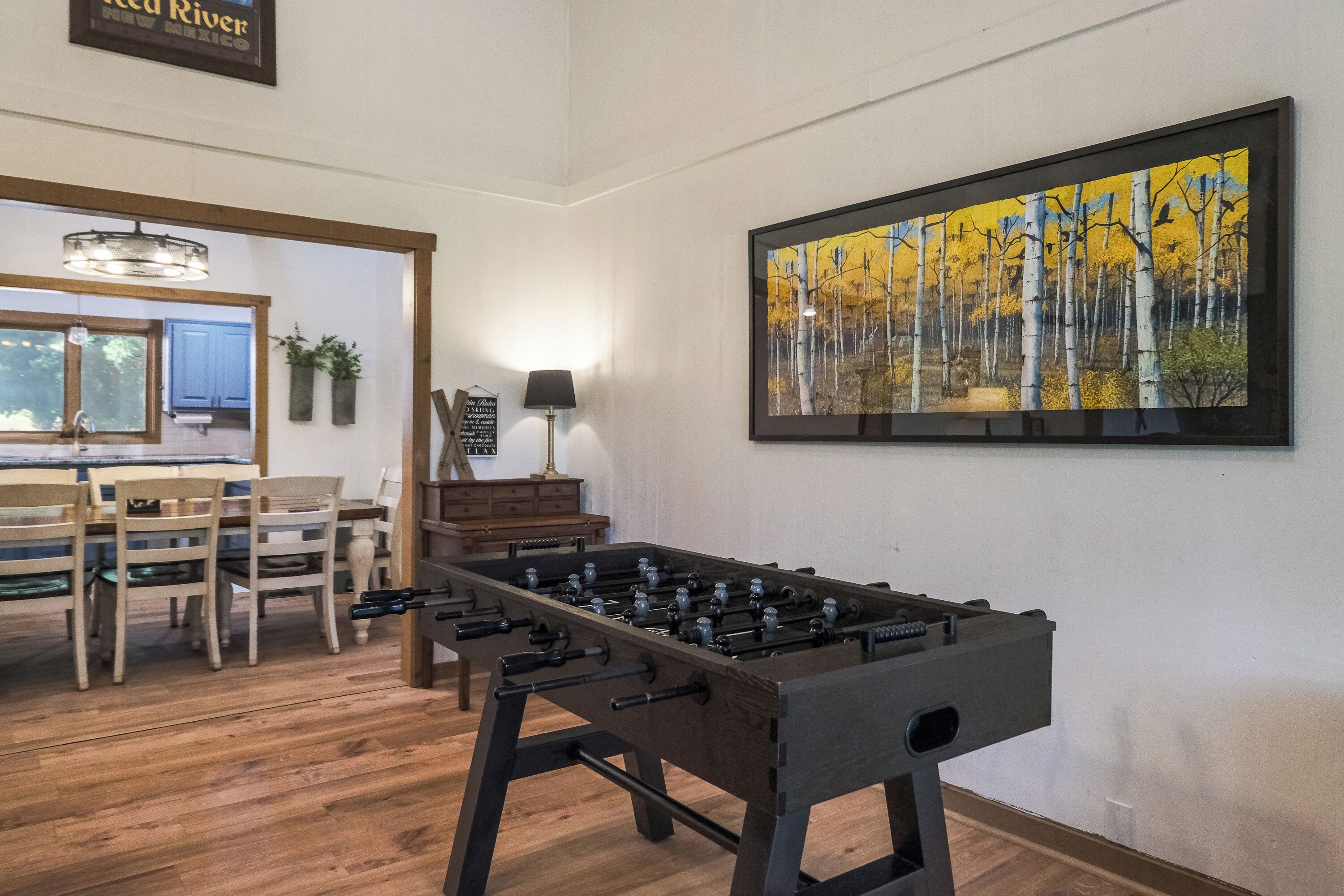 A room with a foosball table, wooden flooring, a dining area with chairs and a table, and a framed painting on the wall.
