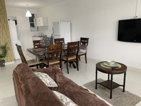 Een moderne woonkamer met een bruine bank, een eettafel met stoelen, een klein rond tafeltje met een schaal en een afstandsbediening, en een televisie aan de muur.