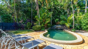 Un patio trasero con piscina, rodeado de abundante vegetación y una terraza de madera con sillones de descanso.
