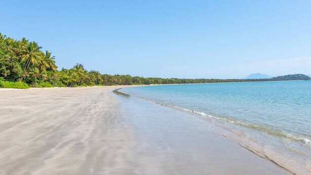 Una amplia playa de arena con agua azul cristalina, palmeras y una montaña distante.