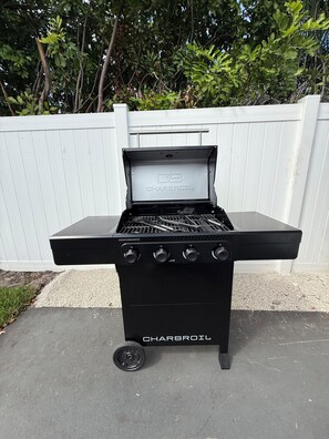 Een zwarte Charbroil buitenbarbecue met vier branders en een bedieningspaneel, geplaatst op een betonnen oppervlak met een witte hek en begroeiing op de achtergrond.