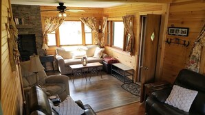 Een gezellig cabin interieur met een stenen open haard, houten wanden, een leren bank, een eethoek en een groot raam met gordijnen.