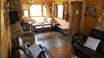 Un accogliente interno di una baita con camino in pietra, pareti in legno, divano in pelle, zona pranzo e un'ampia finestra con tende.