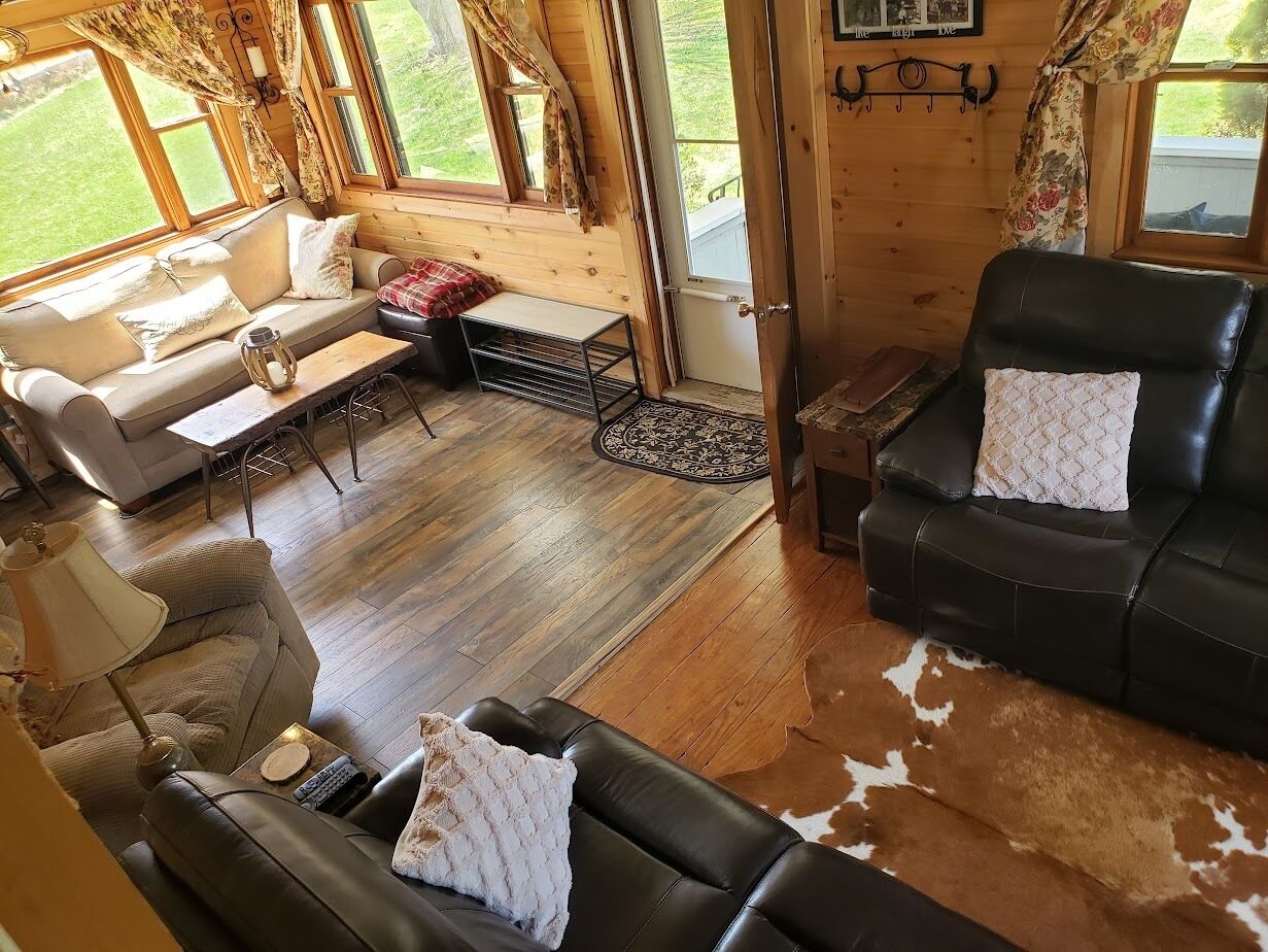 A cozy cabin interior with a sofa, a black leather chair, a wooden floor, and a large window.