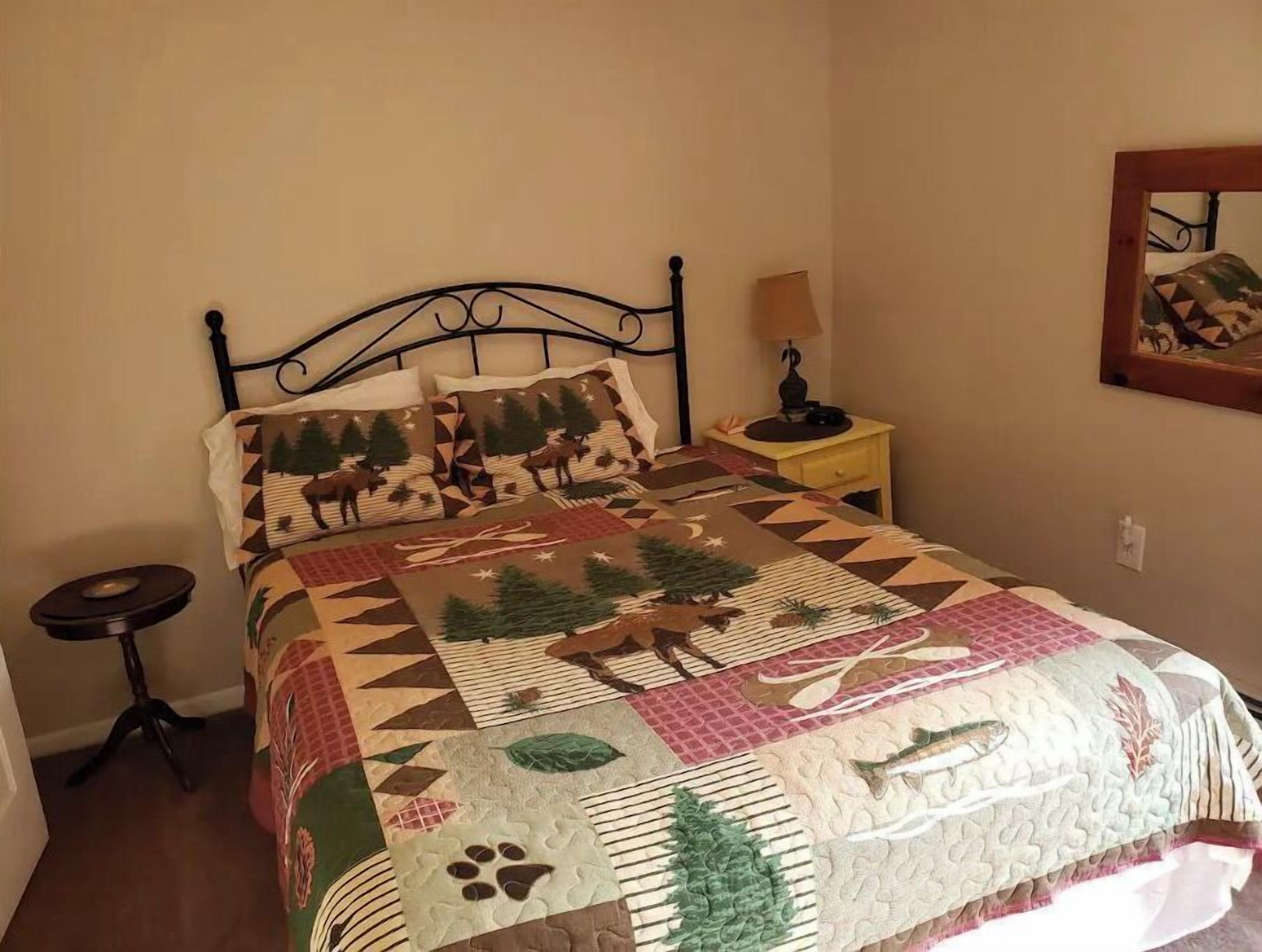 A bed with a geometric patterned quilt, a bedside table with a lamp, and a wall-mounted mirror.