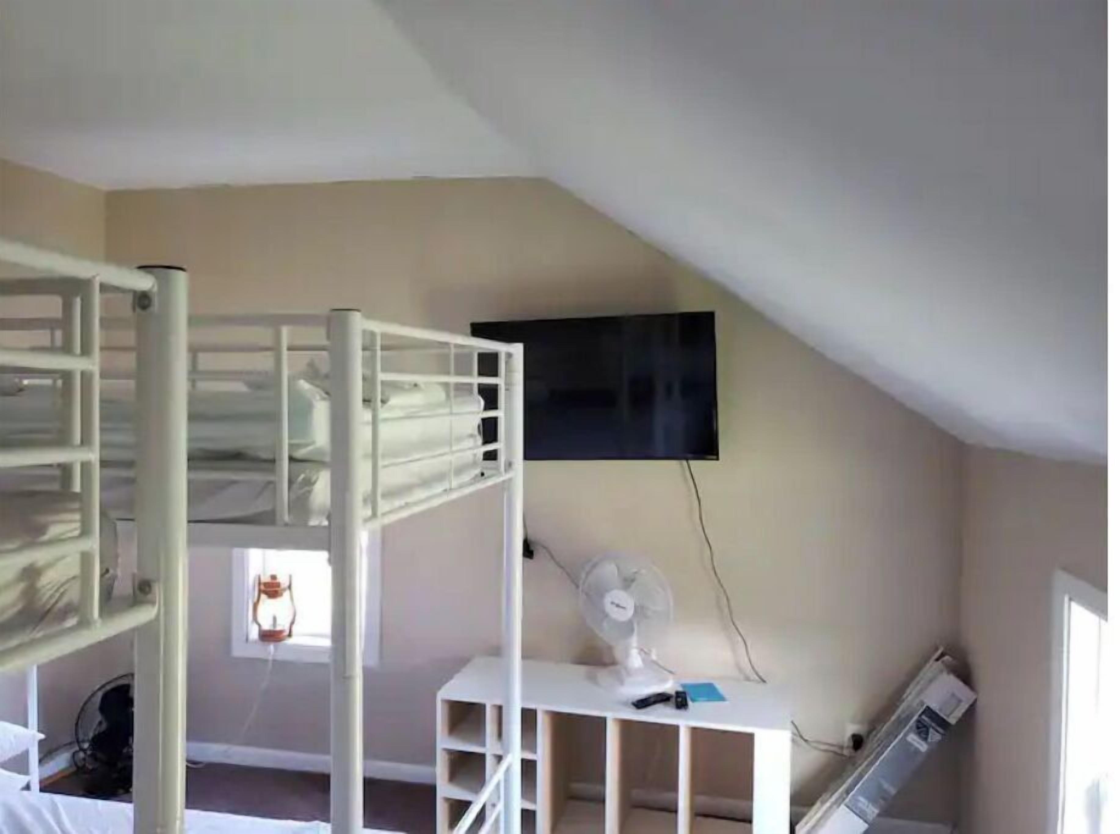 A room with a bunk bed, a desk with a fan, a television mounted on the wall, and a window.