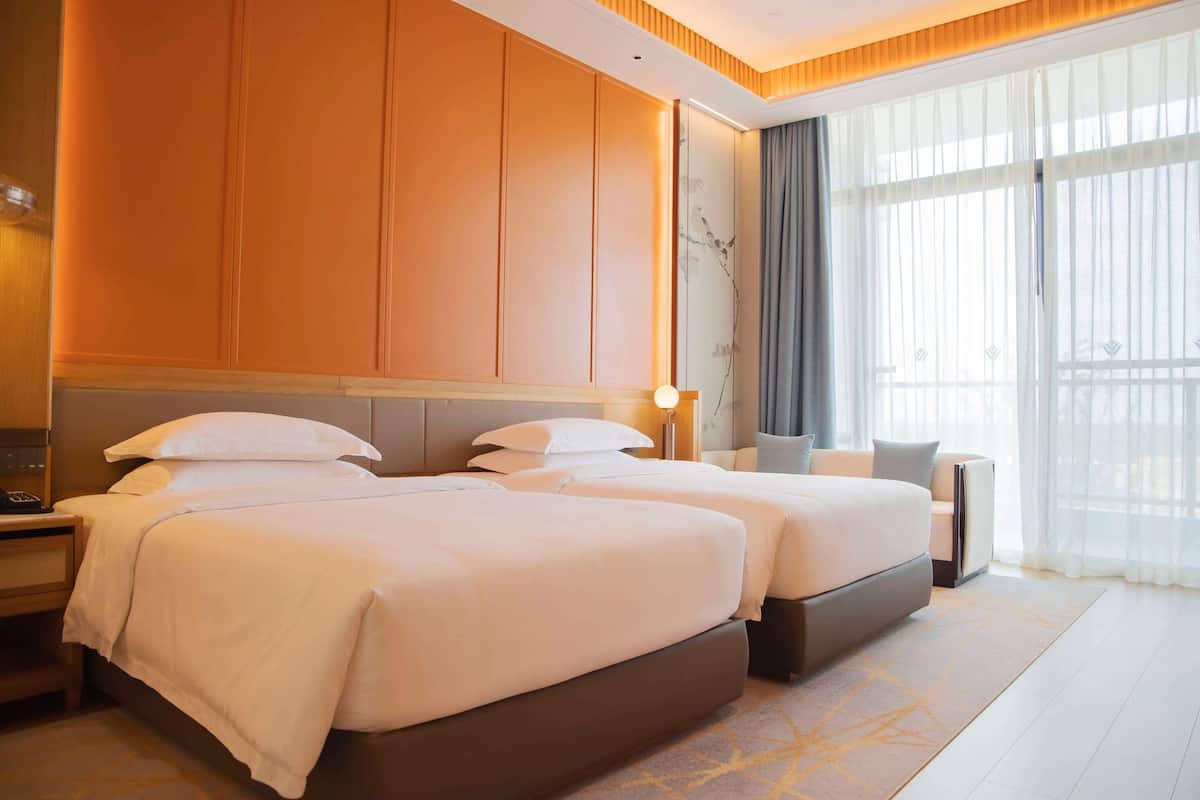 Ein Hotelzimmer mit zwei Betten, einem Nachttisch, einer Lampe und einem großen Fenster mit Vorhängen.